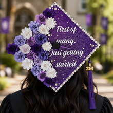 Load image into Gallery viewer, A decorated purple glitter graduation cap worn by a person outdoors on a blurred campus background. The cap features white and purple fabric flowers, a beaded border, white script text reading 'First of many, Just getting started,' a gold '2024' charm, and a purple tassel.