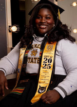Load image into Gallery viewer, Teekajame Creations custom satin graduation stole in gold and black worn by a smiling graduate in a cap and gown.
