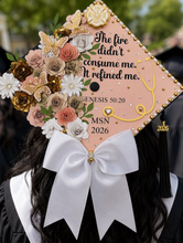 Load image into Gallery viewer, A close-up of a decorated graduation cap worn by a person in a black and white graduation gown. The cap is peach-colored with a large white bow, gold embellishments, and a floral arrangement in peach, white, and gold.
